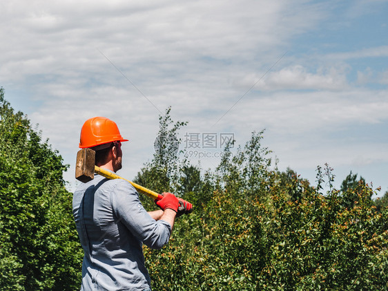 在绿树背景下有吸引力的男子在公园中握着大锤子关闭劳动和就业概念在公园中握着锤子的人衬衫们橙图片