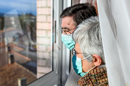 戴防护面罩的老年夫妇在家看窗社交距离封锁老人和退休的生活方式新冠疾团结图片