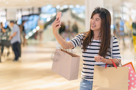 购物曼谷折扣亚洲妇女步行使用智能手机自拍模特通过商店面的眼镜展示装饰服的模型在部门中心欢乐地行动起来亚洲妇女走路用智能移动电话自图片