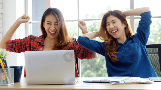 两位年轻女使用笔记本电脑手持微笑地脸在办公桌工作时HappyAsiia女孩完成了项目商业功在线教育家工作的伙伴关系等业务成功项目图片
