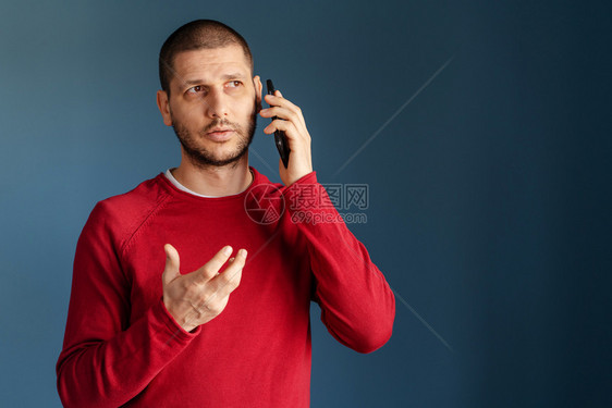 保持聪明的打手势穿着红色毛衣的白人男子站在蓝背景墙前使用智能手机打来电话用手牵说穿着红色毛衣图片