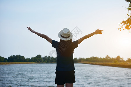湖阳光夏天那女人穿着白色T恤衫和帽子站在河边双手天空上图片