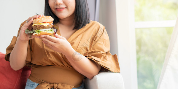 牛肉汉堡饥饿的超重妇女笑着拿汉堡包坐在客厅里她非常快乐并享受吃餐狂食饮紊乱概念小吃坐着图片