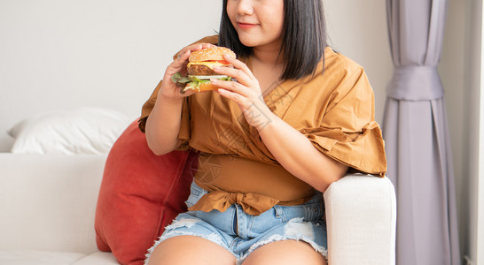 一顿饭高的女孩饥饿超重妇女笑着拿汉堡包坐在客厅里她非常快乐并享受吃餐狂食饮紊乱概念图片