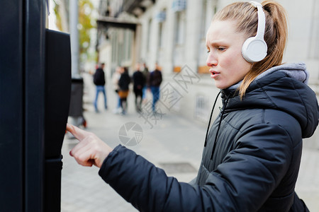 快乐的友好内容年轻金发美女在城里付停车费脸上戴着耳机的夹克脸型惊讶的图片