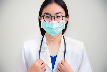 工作室前面肖像对着美丽的亚洲年轻女青医生她带着白制服的听诊器戴眼镜和绿色面具以保护Corona的健康保护Corona在灰色背景上图片