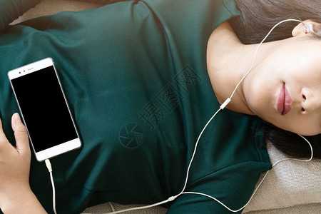 放松好的请享用在沙发上躺下用智能手机听音乐的美丽年轻女头部一眼图片