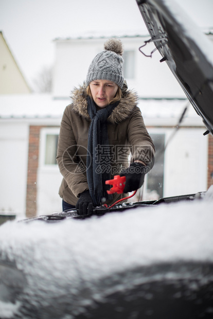 帮助破碎的保险雪天在汽车电池上使用跳跃缆的妇女图片