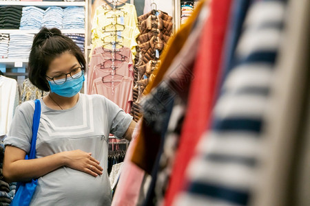 爆发预防亚洲孕妇在购物中心服装店选择新衣时带着面罩保护Corona的亚洲孕妇同时在商店服装中选择新的服装饰精品店图片