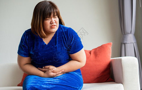 发牢骚痛便秘超重的亚洲妇女坐在客厅沙发上由于胃炎和保健概念手抓着肚子b由于胃部不适症和健康护理概念背景图片