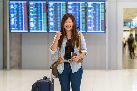 白种人在现代机场飞行信息屏幕上登机旅行和带有技术概念的交通方面携带行李和护照的亚洲旅行者在飞板上走过机牌在行信息屏幕上报到达电话图片