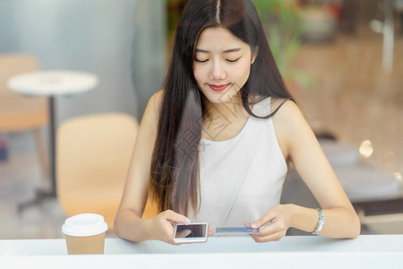 使用带手机的信卡在咖啡店或办公场所在线购物的亚洲年轻妇女除了窗户镜子技术资金钱包和网上支付概念信用卡模型外还信卡和手机在咖啡店或图片