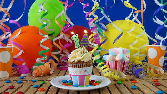 惊喜快乐的老生日14小纸杯蛋糕在质朴的木桌上燃烧着蜡烛背景是五颜六色的气球塑料杯和糖果背景是蓝色的派对墙背景图片