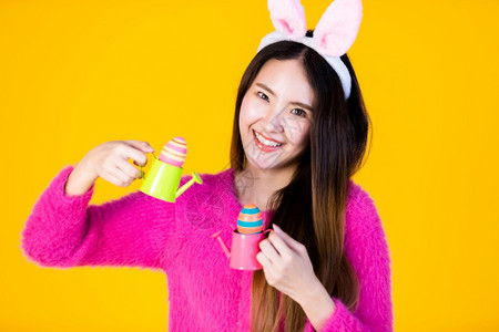 欢笑的亚洲年轻女身着兔子耳朵和小水罐与多彩的复活节鸡蛋相配侧眼看着机在黄色空白副本上隔绝木制的微笑庆典图片