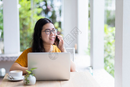 在线的抓住快乐亚洲自由职业者商务女拿着智能手机休闲使用笔记本电脑和咖啡杯在店的背景通信概念无线的图片