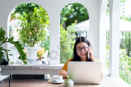 互联网杯子工作区快乐的亚洲自由职业者商务女拿着智能手机休闲使用笔记本电脑和咖啡杯在店的背景通信概念图片