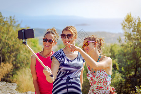 三个穿暑期服装的漂亮女孩旅行者秋天在海上山外用自相棍友情用智能手机拍自人们快乐连衣裙图片