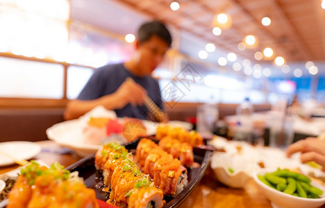人有选择地关注日本餐馆的食本餐厅沙门寿司在一块盘子上有日本食品而餐盘上的模糊男子在餐厅健康亚洲食品沙门寿司菜单中用筷子吃日本食品图片