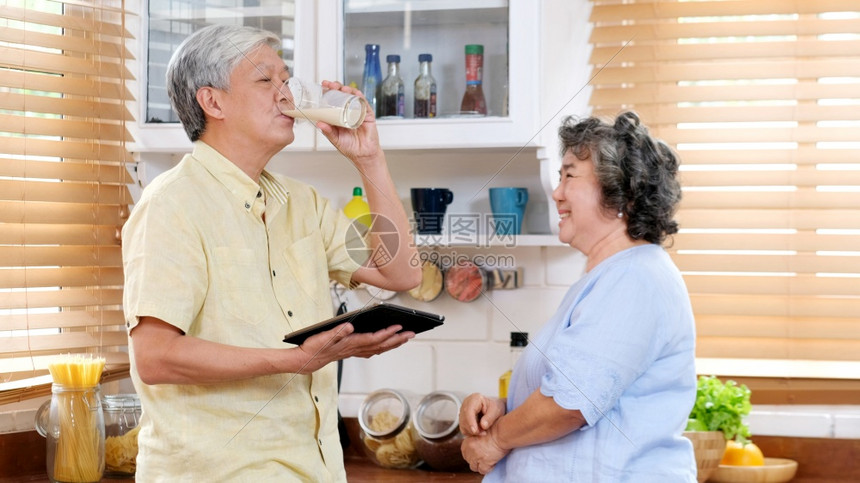 为了高级的家中老年夫妇在厨房里与老年妇女站在一起喝牛奶的老阿西亚男子与妻一起为健康乳奶品拿着牛杯的老年阿西亚男子退休者健康的生活图片
