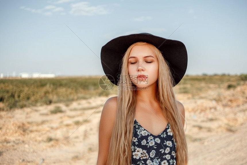 太阳一位年轻美丽的caucasian金发女孩的肖像她穿着蓝色裙子印花粉和黑帽子站在沙乡村公路上夏天日落时靠近葡萄田优雅白种人图片