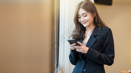 放松在现代办公室或工作场所以及共同工作空间商业生活方式概念的窗口旁边站立着微笑动作的智能手机上使用移动电话对正式西装亚洲女商人的图片