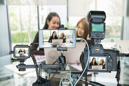 聪明的照片为了两个亚洲年轻女商人通过LiveRiveShalm进行访谈时使用智能移动电话和三脚架行动摄像机的专业照相组企业家实况图片