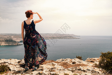 从悬崖起长着裙随风而动手举过头顶看着海边远方的红发年轻女人郎一种冒险图片