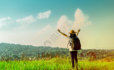 女爬坡道带着帽子和背包站的快乐女游客在山上举起手来在阳光日照耀的子用绿草地举起手来用蓝天和积雪云彩旅行者享受美丽的风景户外图片