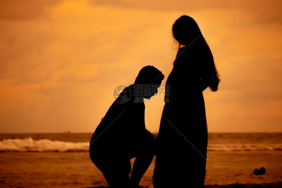 甜的浪漫热情丈夫在海滩上跪下晚日落的休眠亲吻怀孕的老妇人肚子做父母的概念图片