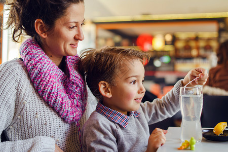 微笑三岁的母亲和儿子坐在餐厅桌旁或咖啡馆儿童边的年幼女子在准备饮料时拿着塑勺或稻草一边喝着欢乐3岁的母亲和儿子年轻女人一边坐在饭图片