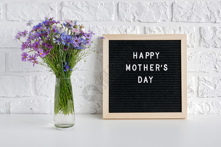 黑色信板上的母亲节快乐文本和桌上花瓶中的蓝色束靠在白砖墙上贺卡明信片的母亲节模板字植物优质的图片