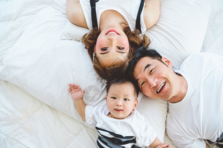 婴儿定型枕在家卧室的白枕床上父亲和母及儿子坐在白枕头床上的亚洲家庭最佳景象快乐的父母和婴儿概念与童的概念在Covid19或Corona的检背景