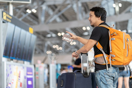 夏天休息室技术在现代机场旅行和运输概念的飞行信息屏幕上登入飞行信息屏幕时对携带行李和护照检查并指向飞行板的亚洲旅者进肖像式监视以图片