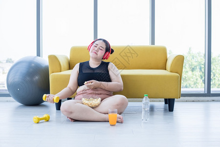 屋合身亚洲超重女使用平板和戴耳机听音乐她在家里运动亚裔女孩享受食物和爆米花的吃量仰卧起坐图片