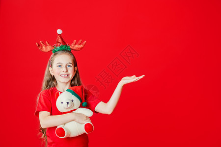 一个有趣的快乐女孩肖像头上缠着角绷带抱一只泰迪熊穿着圣诞睡衣背景为鲜红色头抱着一只泰迪熊与鲜红色背景隔开青春期前打手势戏服图片