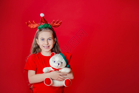 一个有趣的快乐女孩肖像头上缠着角绷带抱一只泰迪熊穿着圣诞睡衣背景为鲜红色头抱着一只泰迪熊与鲜红色背景隔开驯鹿童年笑声图片