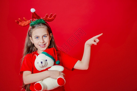 一个有趣的快乐女孩肖像头上缠着角绷带抱一只泰迪熊穿着圣诞睡衣背景为鲜红色头抱着一只泰迪熊与鲜红色背景隔开打手势表明帽子图片