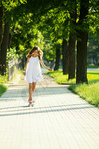 微笑骑术穿着白裙子的可爱城市妇女穿着粉红色溜冰鞋在公园骑着滑板的年轻女孩快乐图片