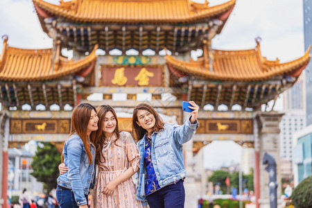 金碧信息在昆明金比广场旅行时使用智能手机自拍带着友谊观的旅行和游概念的3个亚洲幸福妇女一起穿行于昆明市金比广场城景观图片