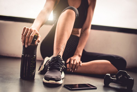 运动员女孩腿近身子在室内健房锻炼运动在体育训练后休息使用哑铃智能电话和蛋白奶昔瓶健康生活方式Athlete和健体肌肉概念图片