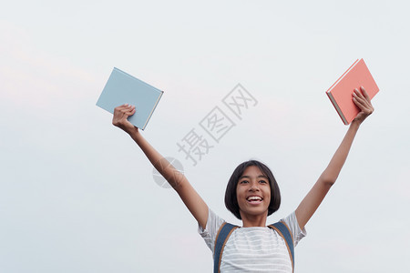 快乐的亚洲女孩在拿着一本书和手举到蓝天背景时脸上微笑容瞳孔学习生图片