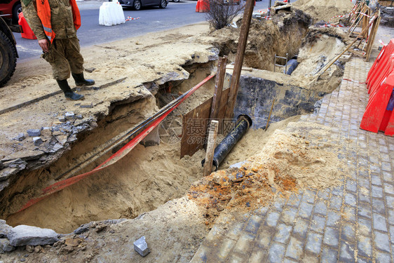 洞铺路危险的一名道服务工人站在公路上挖掘的壕沟附近检查混凝土区水井城市下道管和铺设的泥板修复街道路上的城市下水图片