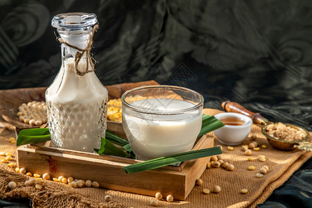 一杯自制豆类牛奶和大装在木盘中用棕色糖和蜂蜜供应晨亮时有照明的替代牛奶概念FrontView选择焦点复制空间种子产品白色的图片