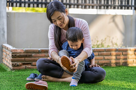 妈快乐的亚洲单身母亲在现代自学或家庭校和单身母亲概念选择重点的现代住房前草坪上为儿子穿亚洲单身母亲鞋活的背景图片