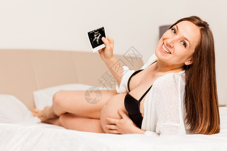 内衣内裤幸福孕妇躺在床上并拿着婴儿扫描图像的相片腹部奢华背景
