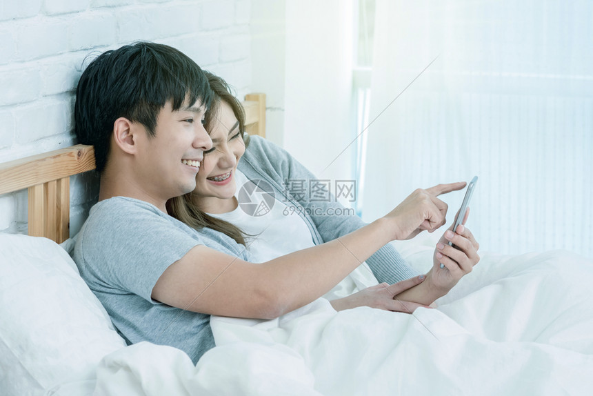 吸引人的房间假期在现代家庭用智能手机在床上的铺用手机情侣和生活方式概念指向男朋友来拉近亚洲快乐情人或夫妇之间的距离图片