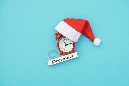 桌子日程小时木制历冬季月份12红色闹钟和蓝纸背景上的圣诞帽平躺图片