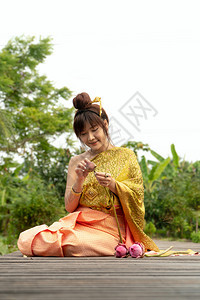 身着传统泰式礼服坐在木桥上的女美丽她手在泰国风格下举手受人尊敬在莲花叶上还有粉红莲女孩快乐的裙子图片