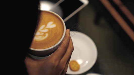 咖啡杯中最顶端的闭紧拿铁艺术由妇女手握在咖啡店或书的杯这些铺具有低光饮料和反组概念假期思维曼谷图片