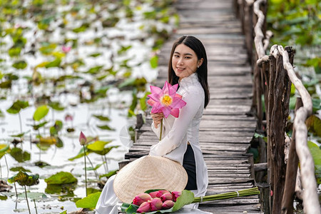 越南女士语在大莲花湖维特南青山或东方的木桥上挂着粉色莲花的传统维特南帽子坐在木桥上的美丽女肖像图片